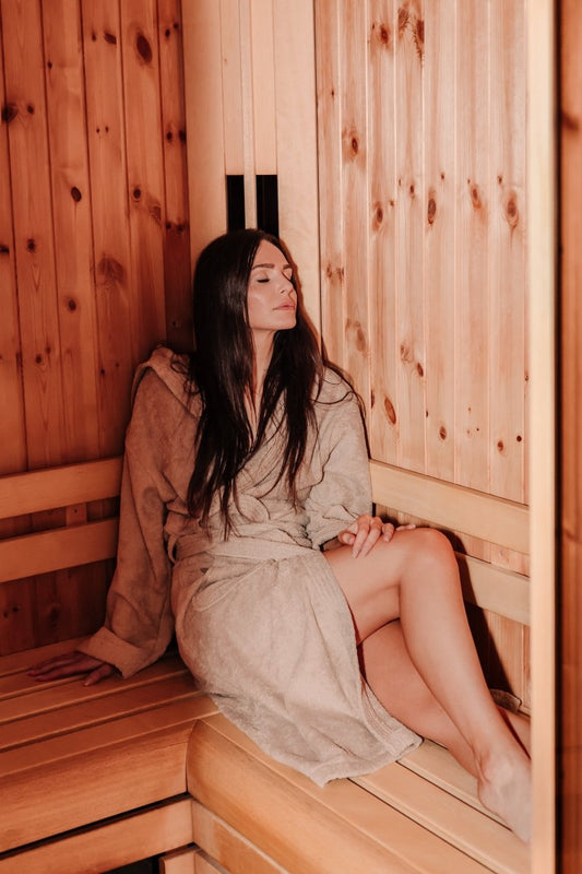Een vrouw zittend in een houten sauna, gekleed in een lichte badjas, met haar haar loshangend en een ontspannen uitdrukking.