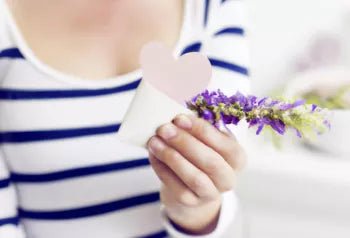 Een close-up van een vrouw in een gestreept shirt die een witte kaart in de vorm van een hart vasthoudt, naast een takje met paarse bloemen, wat suggereert dat ze een Moederdag-geschenk in de vorm van een badjas heeft ontvangen.
