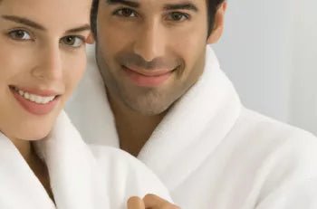 Een man en een vrouw, beide gekleed in witte badjassen, dicht bij elkaar staand en glimlachend naar de camera.